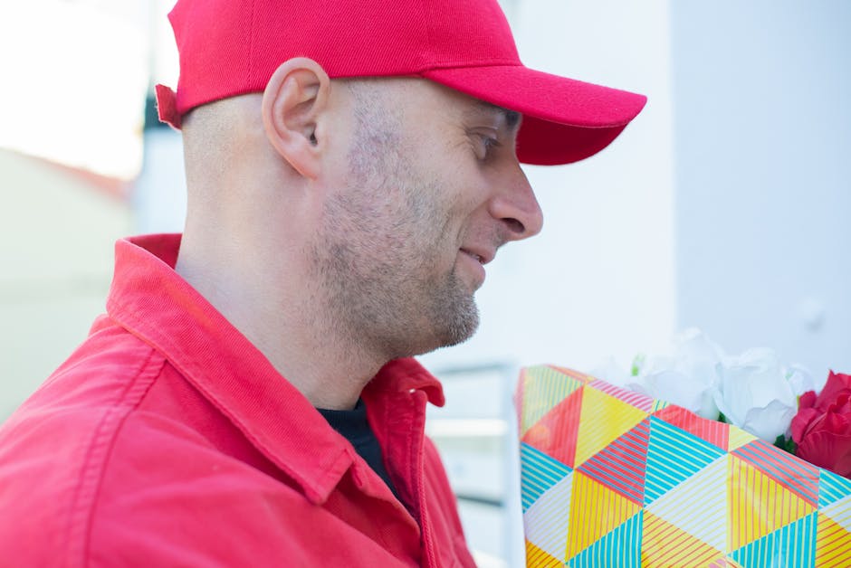 A delivery person wearing a red cap and matching red uniform holding a colorful, geometric-patterned gift bag filled with a bouquet of fresh flowers, including white roses and red blooms, in an outdoo