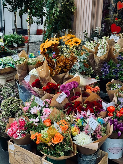 An outdoor display of numerous floral bouquets arranged in metal buckets and wooden crates at eastdulwichflowers.org.uk. The bouquet features a vibrant mix of fresh flowers including pink lilies, oran