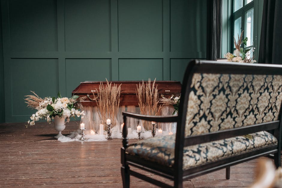 A floral tribute arrangement placed on a wooden table in a softly lit room with green paneled walls and large window, featuring a bouquet of white lilies, roses, and greenery in a white vase, alongsid