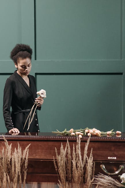 A woman with dark skin and curly hair styled in a high puff, dressed in a black blazer, stands beside a wooden casket adorned with a floral arrangement of pale pink roses and greenery. She holds a sin
