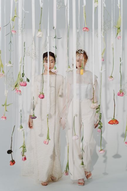 Two women dressed in elegant, light-colored dresses stand behind a delicate curtain of hanging flowers with pink, red, purple, and white blooms, creating a soft floral backdrop. The flowers are suspen